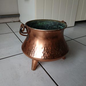 Peut inclure: Un chaudron en cuivre avec trois pieds courts et deux poignées. Le chaudron a une texture martelée sur le corps et une patine vert foncé à l'intérieur. Le pot est en cuivre et est probablement utilisé pour la cuisine ou la décoration.