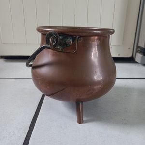 May include: A copper cauldron with three legs and a handle. The pot has a rounded body and a wide rim. The handle is attached to the side of the pot with a metal ring. The cauldron is a reddish-brown color.