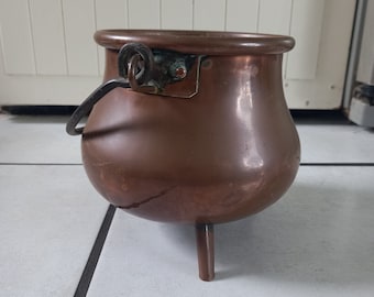 Puede incluir: Un caldero de cobre con tres patas y un asa. La olla tiene un cuerpo redondeado y un borde ancho. El asa está sujeta al lateral de la olla con un anillo metálico. El caldero es de color marrón rojizo.