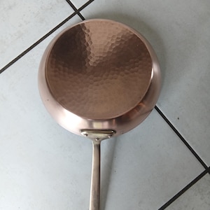 Puede incluir: Una sartén de color cobre con un mango largo. El interior de la sartén tiene una textura martillada. El mango está sujeto con remaches. La sartén está diseñada para cocinar y está hecha de metal.