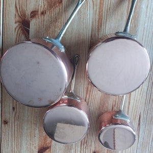 May include: Four copper saucepans with silver handles are arranged against a light brown wooden backdrop. The pans, of varying sizes, have rounded bases and a shiny, reflective surface. Each pan features a loop on the handle for hanging.