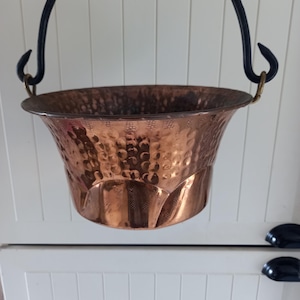 Pode incluir: Um balde de metal martelado de cor cobre com uma alça de metal preta. O balde tem uma borda alargada e uma superfície texturizada. A alça é fixada com ferragens de cor latão. O balde está pendurado em um fundo branco.
