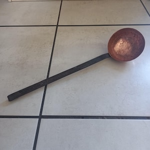 May include: A copper ladle with a long black metal handle. The ladle has a hammered finish and is slightly dented.