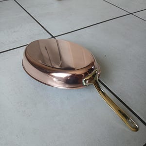 Könnte beinhalten: Eine polierte Kupferpfanne mit einer glatten, reflektierenden Oberfläche und einem langen, goldfarbenen Metallgriff. Die Pfanne hat eine abgerundete, flache Schalenform und ist mit zwei sichtbaren Nieten am Griffansatz befestigt.