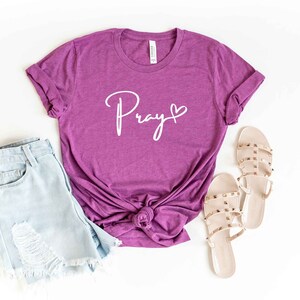 Op de afbeelding: Een paars T-shirt met het woord "Pray" in witte cursieve letters met een klein hartje ernaast. Het shirt is onderaan geknoopt en wordt gecombineerd met lichtblauwe denim shorts en beige sandalen.