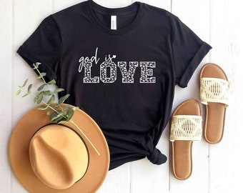 May include: Black t-shirt with the text "God is Love" in white with a leopard print pattern. The shirt is laid flat on a white wooden surface with a brown hat and white sandals.