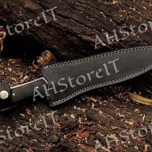 May include: A black leather sheath with a stitched edge and a silver blade knife with a black handle. The knife is on a wooden surface.