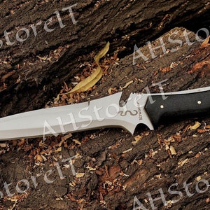 May include: A silver and black knife with a black handle and a snake design on the blade. The knife is resting on a piece of wood.