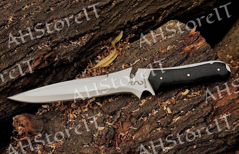 May include: A silver and black knife with a black handle and a snake design on the blade. The knife is resting on a piece of wood.