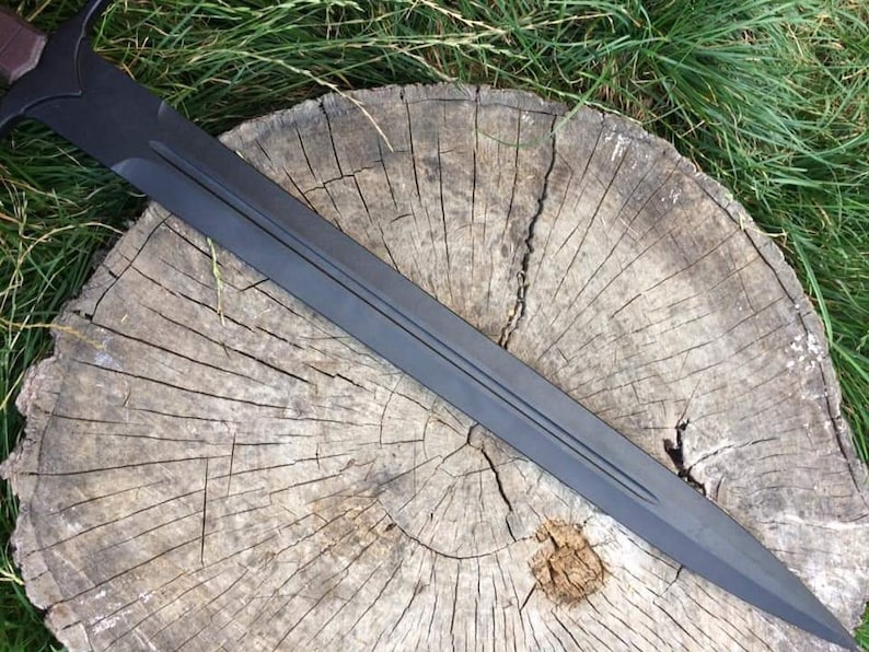 May include: A black sword with a brown handle, lying on a wooden stump. The sword has a double-edged blade and a pointed tip.