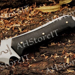 May include: A black handled knife with a silver blade. The blade has a serrated edge and a small hole near the handle. The handle is made of wood and has a brass pin. The knife has the word "Krauser" engraved on the blade.