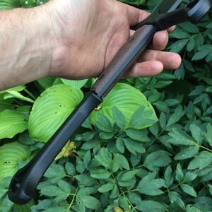 May include: A black and brown sword with a wooden handle. The sword is held in a hand, with the blade pointing towards the camera.