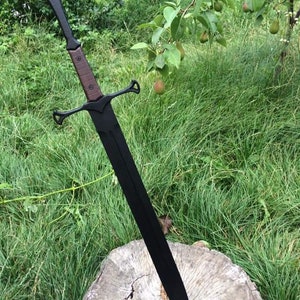 May include: A black sword with a brown handle and a decorative guard. The sword is resting on a tree stump in a grassy area.