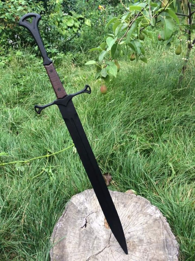 May include: A black sword with a brown handle and a decorative guard. The sword is resting on a tree stump in a grassy area.