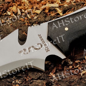 May include: Close-up of a silver knife with a black handle. The knife has a serrated blade and a small hole in the blade. The knife is engraved with the word "Krauser".