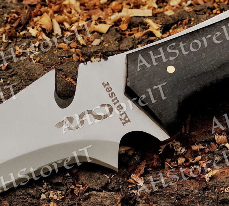 May include: Close-up of a silver knife with a black handle. The knife has a serrated blade and a small hole in the blade. The knife is engraved with the word "Krauser".