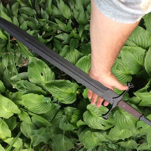 May include: A black sword with a long, straight blade and a decorative hilt. The sword is held by a hand in a garden setting.