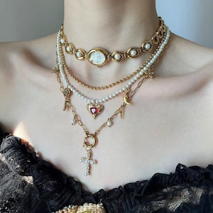 Puede incluir: Conjunto de collares dorados en capas con un reloj, perlas y varios dijes. El collar incluye un colgante de reloj, hilos de perlas, un colgante de corazón con una piedra roja y un colgante de cruz. Los dijes incluyen una llave, una luna creciente y rayos.