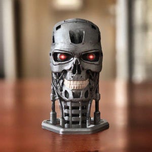 May include: A metallic gray skull-shaped sculpture with glowing red eyes. The sculpture has a detailed skeletal design, including teeth and mechanical components. It sits on a small, geometric base, and is a unique piece of art.
