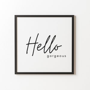May include: A framed print with a white background and black text. The word "Hello" is written in a cursive font above the word "gorgeous" in a smaller, sans-serif font. The frame is black.