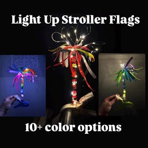 May include: Light Up Stroller Flags with 10+ color options. The product features a vertical pole decorated with colorful beads and illuminated by integrated LED fairy lights, topped with a cluster of vibrant ribbons and a sturdy plastic clamp base.