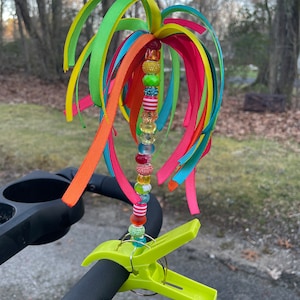 May include: A colorful stroller toy with a beaded string and multiple ribbons in shades of green, yellow, orange, red, and blue. The toy is attached to a stroller with a green clip.
