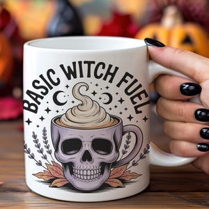 Może przedstawiać: Biały ceramiczny kubek z napisem "BASIC WITCH FUEL". Kubek ma wzór czaszki z filiżanką kawy i bitą śmietaną. Kubek jest trzymany przez osobę z czarnym lakierem do paznokci. Dynie i jesienne liście w tle.