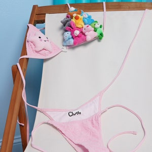 May include: Pink bikini with a star-shaped top and a thong bottom. The word "Clush" is printed on the inside of the bottom. A collection of small plush toys in various colors is suspended above the bikini.