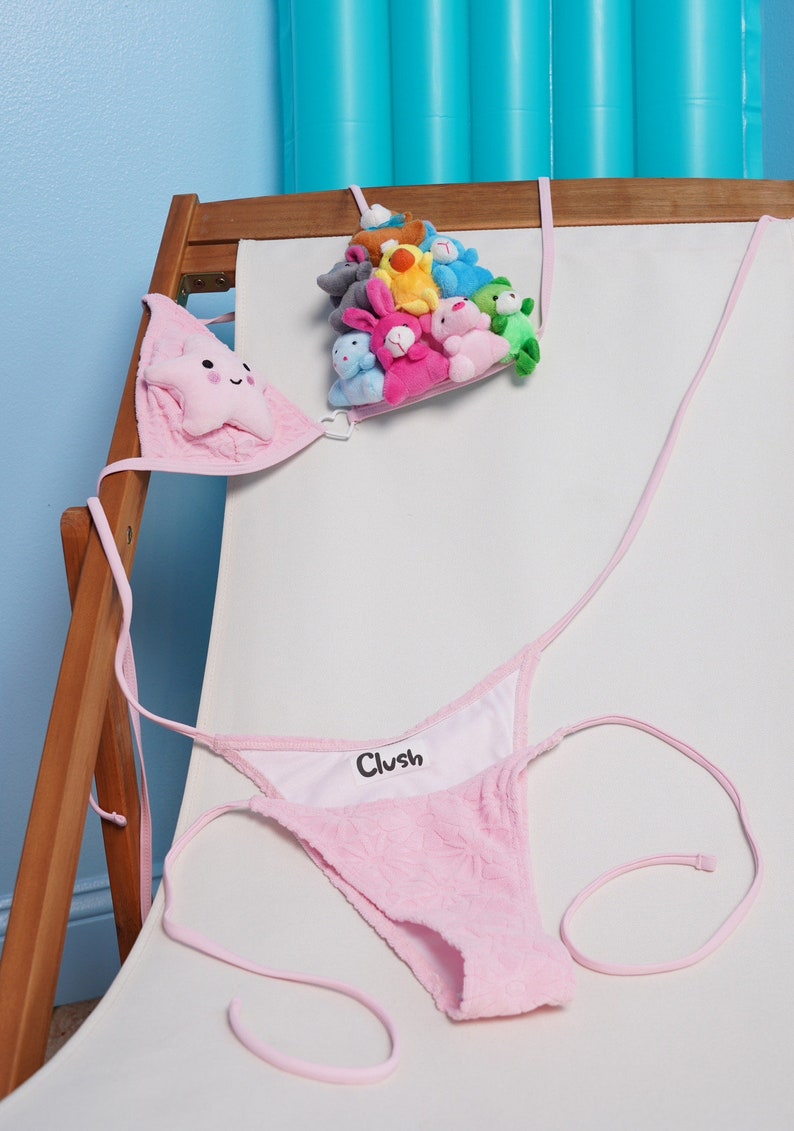 May include: Pink bikini with a star-shaped top and a thong bottom. The word "Clush" is printed on the inside of the bottom. A collection of small plush toys in various colors is suspended above the bikini.