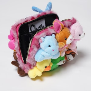 May include: A pink plush zippered pouch with a black zipper and a black fabric interior. The pouch is decorated with a variety of colorful plush animal toys, including a blue hippo, a yellow chick, a green frog, an orange bunny, and a pink pig. The pouch has a label that reads "Clutch".