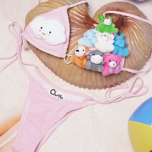 May include: Pink bikini set with a cloud-shaped appliqu&eacute; on the top. The top and bottom are made of a textured fabric. The top features a smiling cloud and the bottom has the word "Clush". Small stuffed animals are on the seashell.