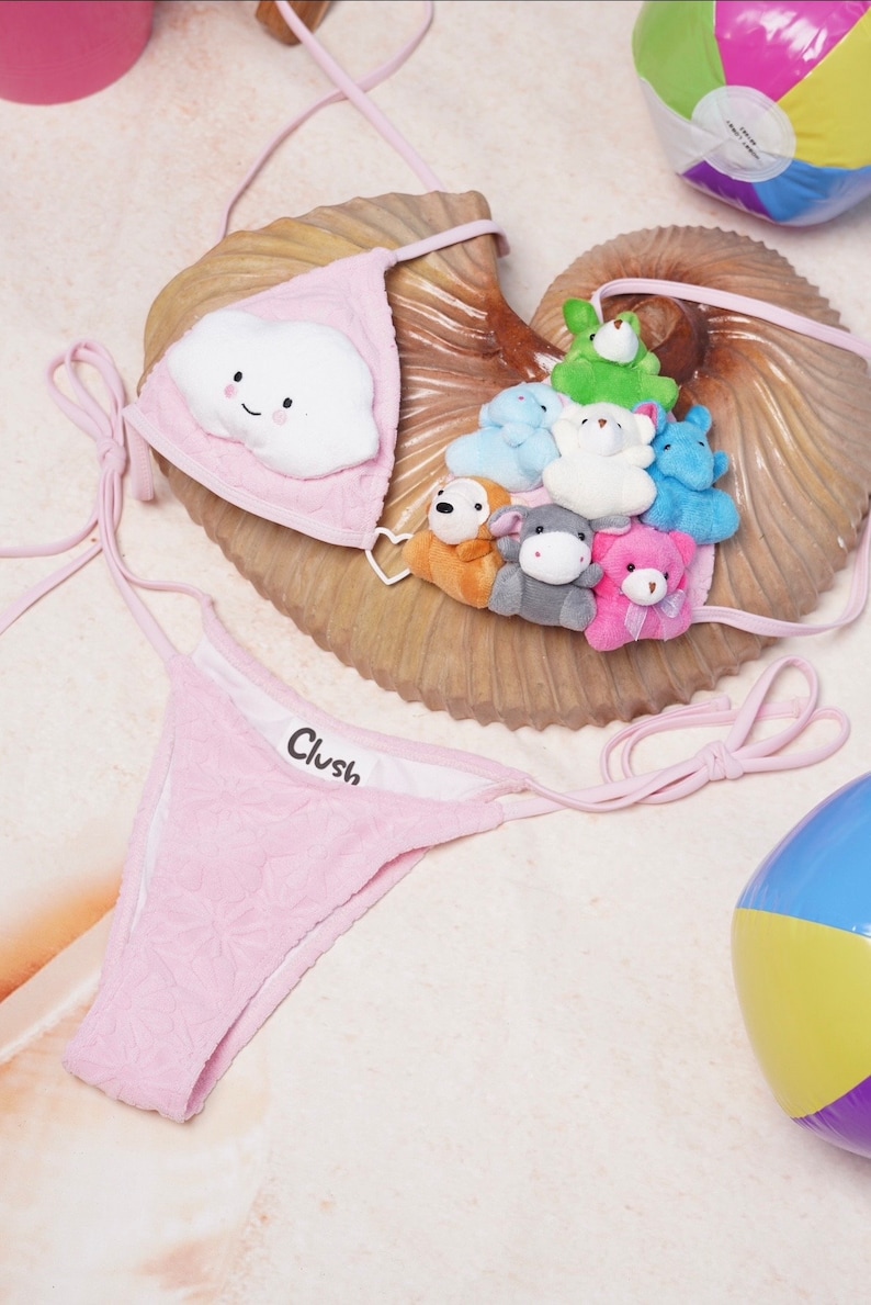 May include: Pink bikini set with a cloud-shaped appliqu&eacute; on the top. The top and bottom are made of a textured fabric. The top features a smiling cloud and the bottom has the word "Clush". Small stuffed animals are on the seashell.