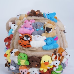 Puede incluir: Una mochila beige cubierta de varios juguetes de animales de peluche. La mochila tiene un compartimento principal con cremallera y está adornada con numerosos animales de peluche pequeños, incluyendo un león, un tigre y un pato. Los peluches son de varios colores y tamaños.
