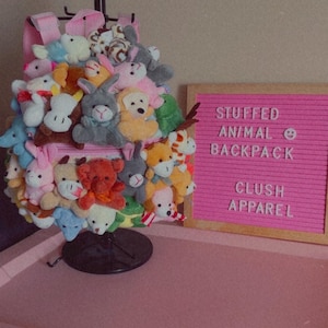 May include: A pink stuffed animal backpack filled with various plush toys. The backpack is on a black stand and a pink letter board behind it reads "STUFFED ANIMAL BACKPACK" and "CLUSH APPAREL".