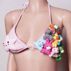 May include: Pink bikini top with a star-shaped design on one cup and a cluster of plush animal toys on the other. The bikini features a halter neck tie and a heart-shaped connector.
