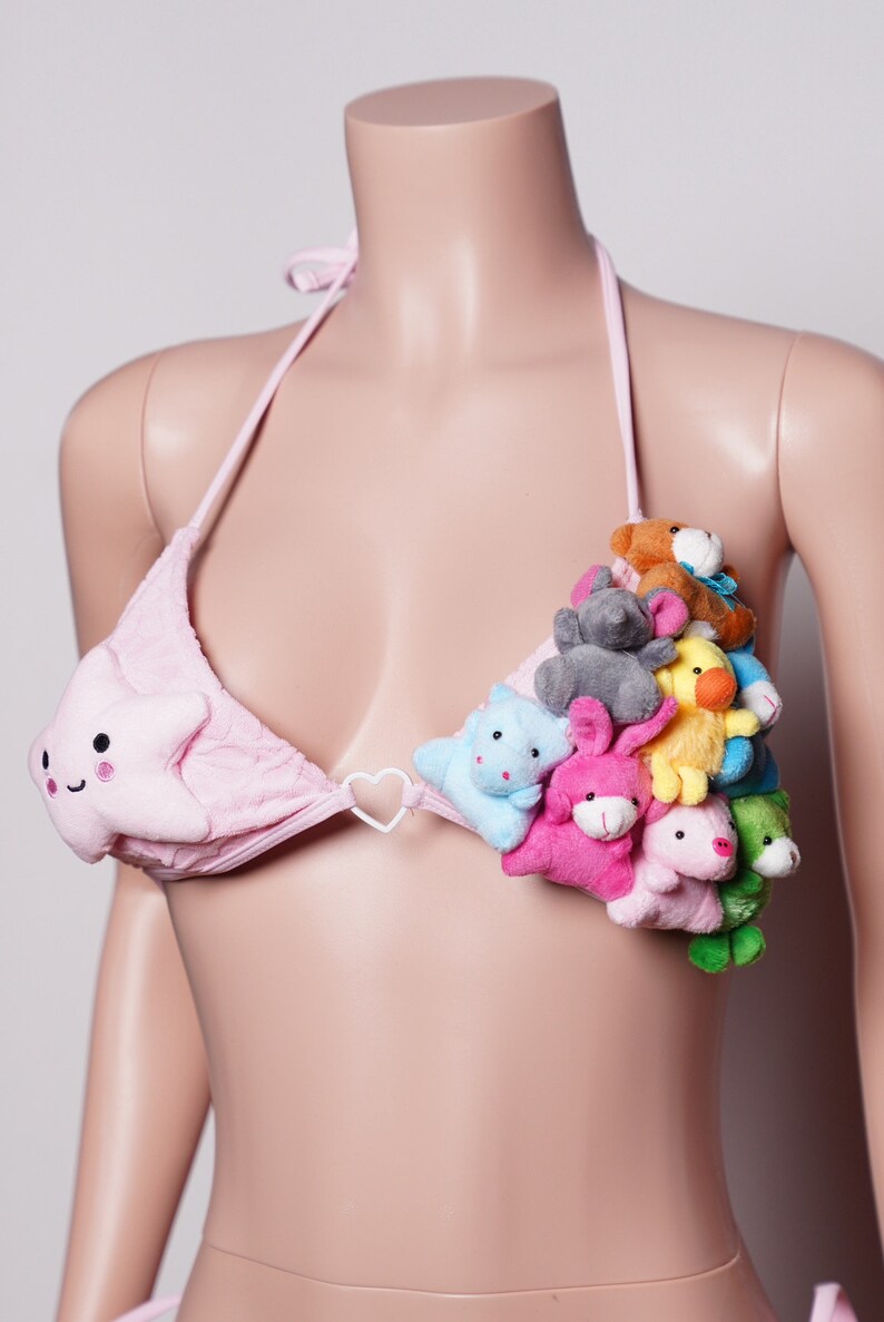 May include: Pink bikini top with a star-shaped design on one cup and a cluster of plush animal toys on the other. The bikini features a halter neck tie and a heart-shaped connector.