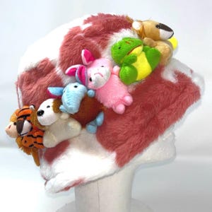 Puede incluir: Un sombrero caprichoso con un estampado de lunares rojos y blancos, adornado con una variedad de pequeños animales de peluche de colores. Los animales incluyen un tigre, un león, un perro, un elefante, un conejito y una rana.