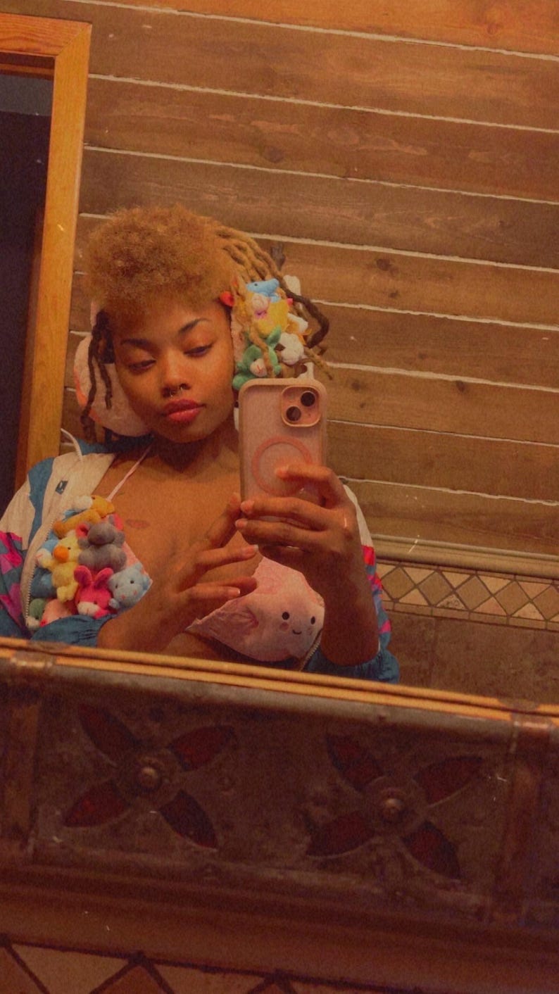 May include: A selfie in a bathroom mirror. The person is wearing a jacket and a top adorned with plush toys. The hair is styled with colorful accessories. The phone case is pink. The mirror frame has a decorative design.