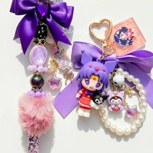 May include: Two decorative pens and a keychain featuring cartoon characters. The pen on the left has a black and white character with a purple bow, a pink pom-pom, and a glittery base. The keychain has a heart-shaped clasp and a character with a purple bow.