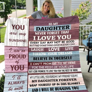 May include: A pink and blue blanket with a woodgrain background. The blanket has a message for a daughter from her dad, saying "To my daughter, never forget that I love you." The blanket also includes the words "laugh, love, live, follow your dreams, believe in yourself, and love, dad."