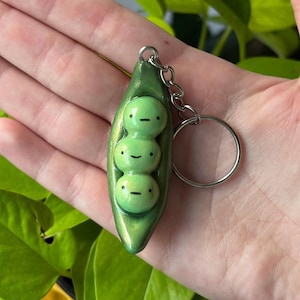 May include: A green pea pod keychain with three smiling peas inside. The keychain is made of a shiny green material and has a silver keyring attached. The peas have small black eyes and mouths.