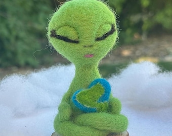 Puede incluir: Una figurita de extraterrestre de fieltro verde con los ojos cerrados, labios rosados y un corazón azul y verde en sus manos, sentada en una postura meditativa sobre una base de madera.