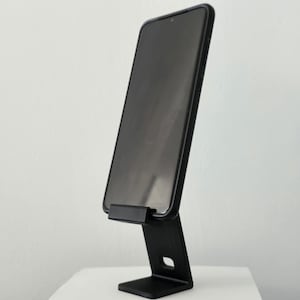Könnte beinhalten: Ein schwarzer Smartphone-Ständer mit einem Telefon im Hochformat. Der Ständer ist aus Metall und hat ein schlichtes Design.