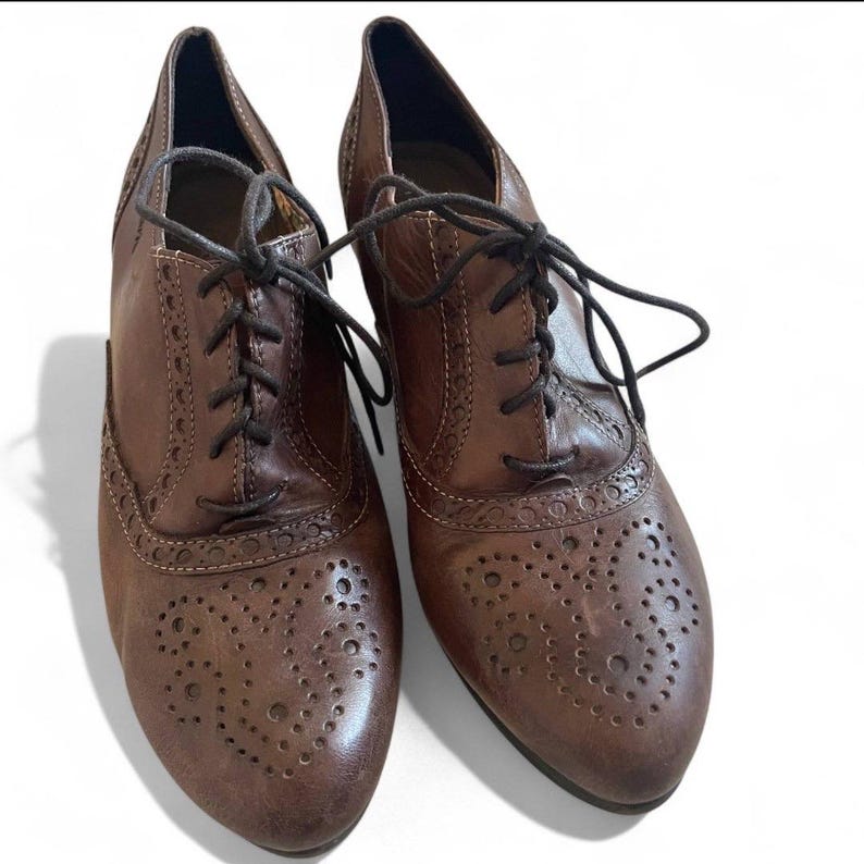 May include: A pair of brown leather brogue shoes with a lace-up design. The shoes have decorative perforations on the toe and sides, and a small heel. The leather has a distressed appearance.