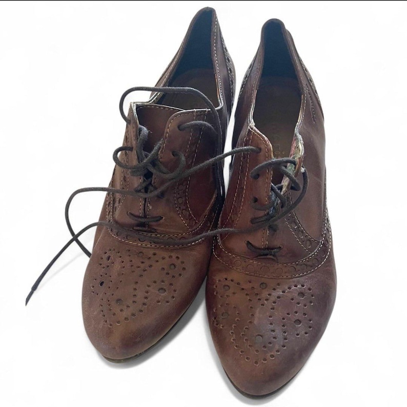May include: A pair of brown leather heeled oxfords with lace-up closures. The shoes have decorative perforations on the toe and sides. The heels are approximately 7.6 cm high.