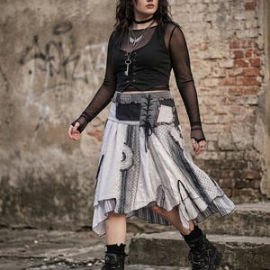 以下が含まれることがあります： 黒いメッシュの長袖トップス、黒いベスト、グレーと白の色合いのパッチワークスカートを着た女性。厚底とストラップが付いた黒いブーツを履いています。この服装はゴシックまたはオルタナティブスタイルです。