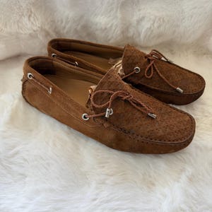 May include: A pair of brown suede driving moccasins with leather laces and silver-tone eyelets. The shoes have a textured surface and are displayed on a white, fluffy background.