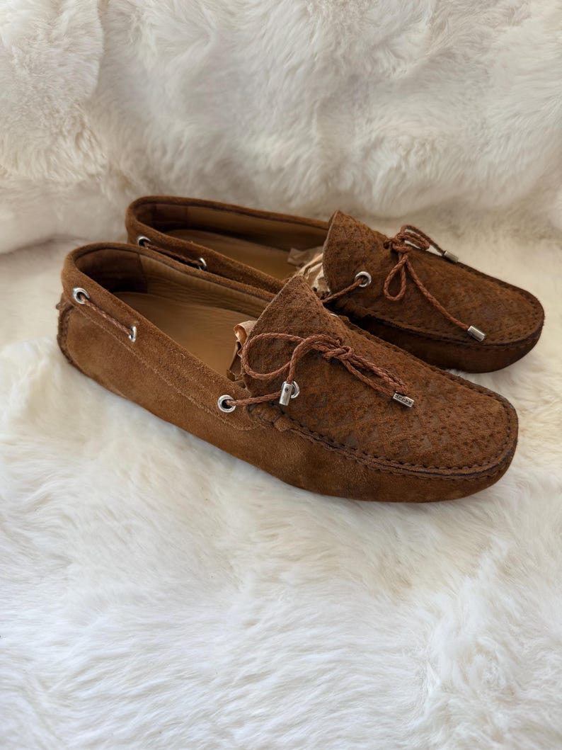 May include: A pair of brown suede driving moccasins with leather laces and silver-tone eyelets. The shoes have a textured surface and are displayed on a white, fluffy background.