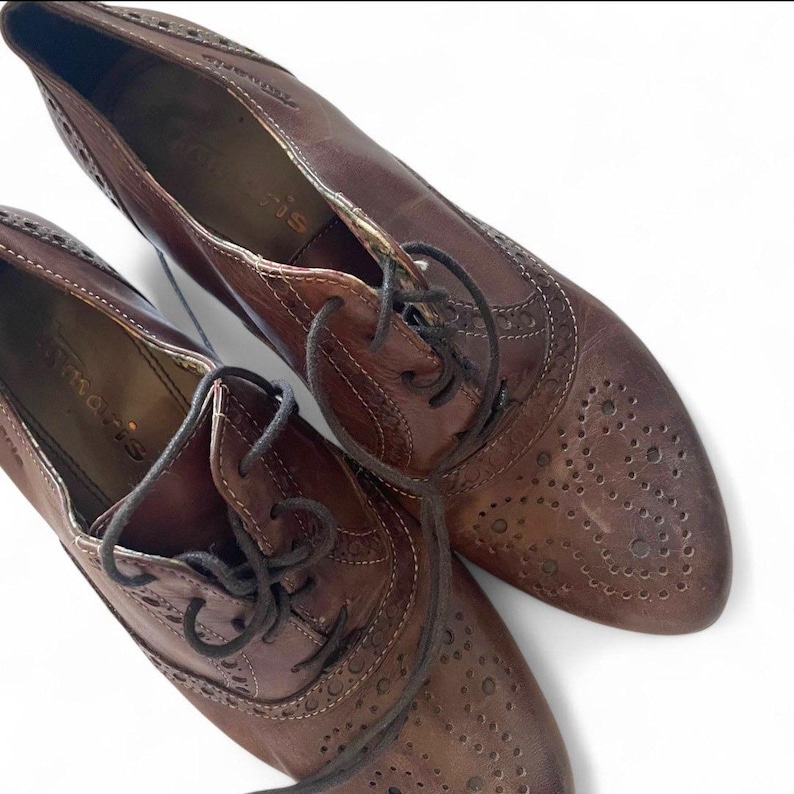May include: A pair of brown leather oxford shoes with decorative perforations and laces. The shoes have a classic design with a pointed toe and a low heel. The brand name "Tamaris" is visible on the inner sole.
