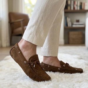 May include: Brown suede driving moccasins with a textured pattern and a leather tie detail. The shoes are worn with cream-colored pants on a white fluffy rug. The shoes have a classic, casual style.